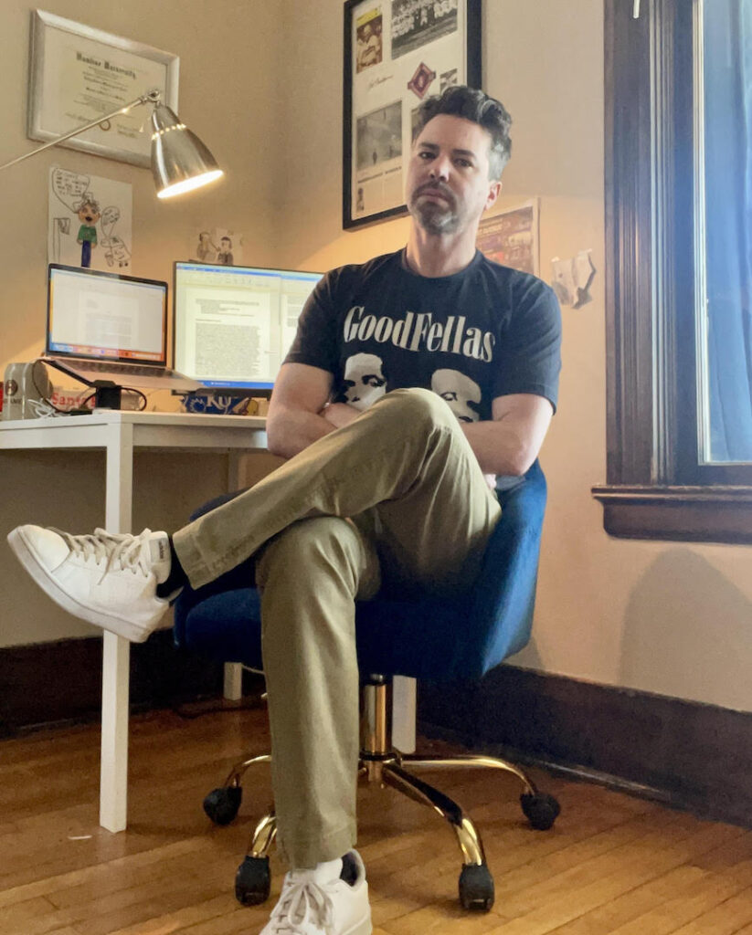 Casey DW Jones, literary fiction author, sits in a blue swivel chair in front of his computer, wearing a Goodfellas t-shirt.